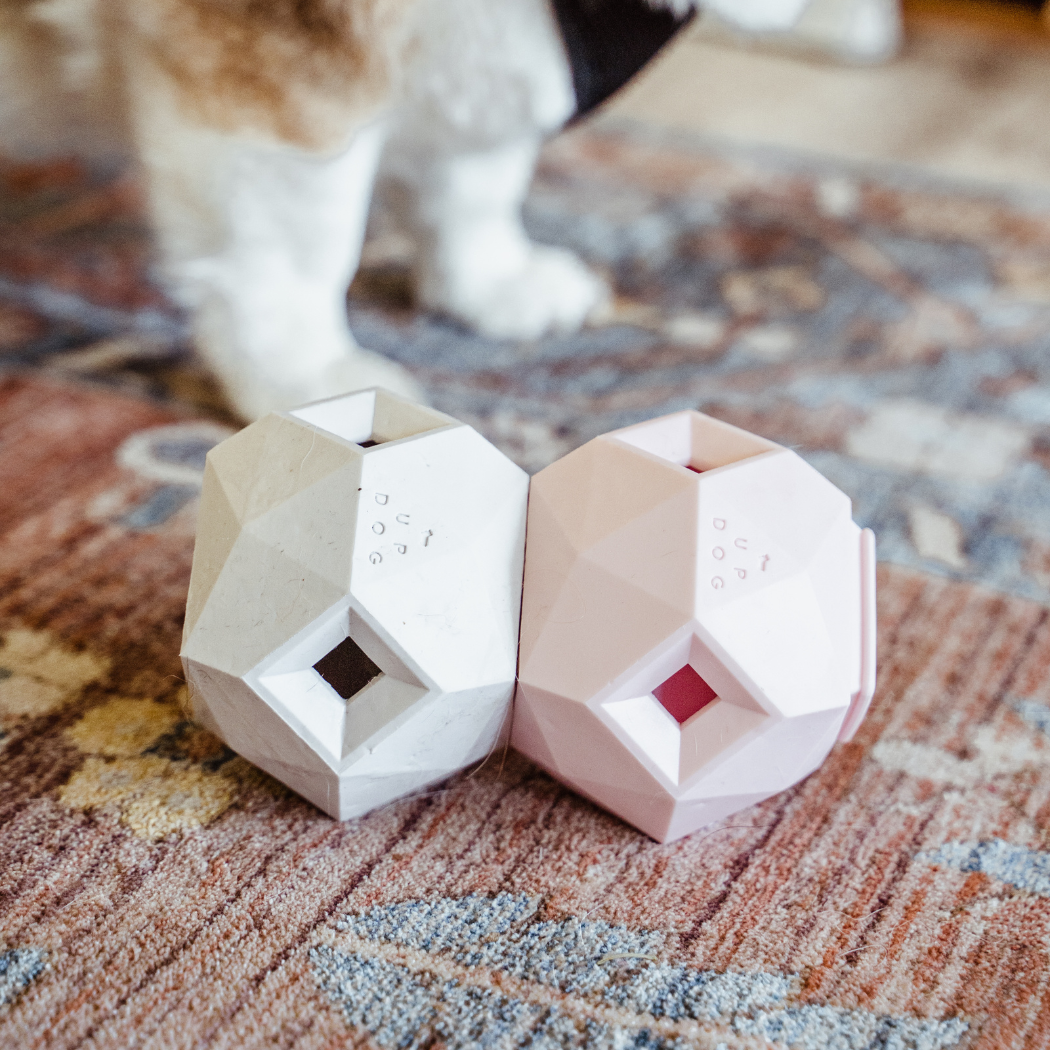 Up Dog Odin Treat Puzzle Balls in light grey and rose quartz, two linked side by side on a patterned rug