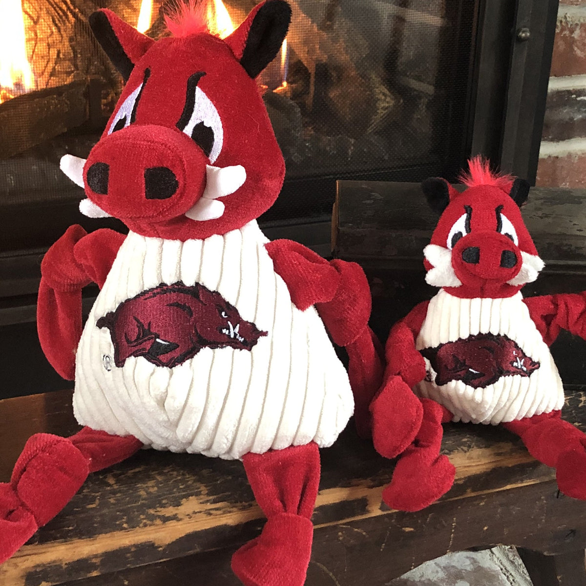 Tusk the Razorback Knottie dog toy in two sizes on a wooden bench in front of a lit fireplace