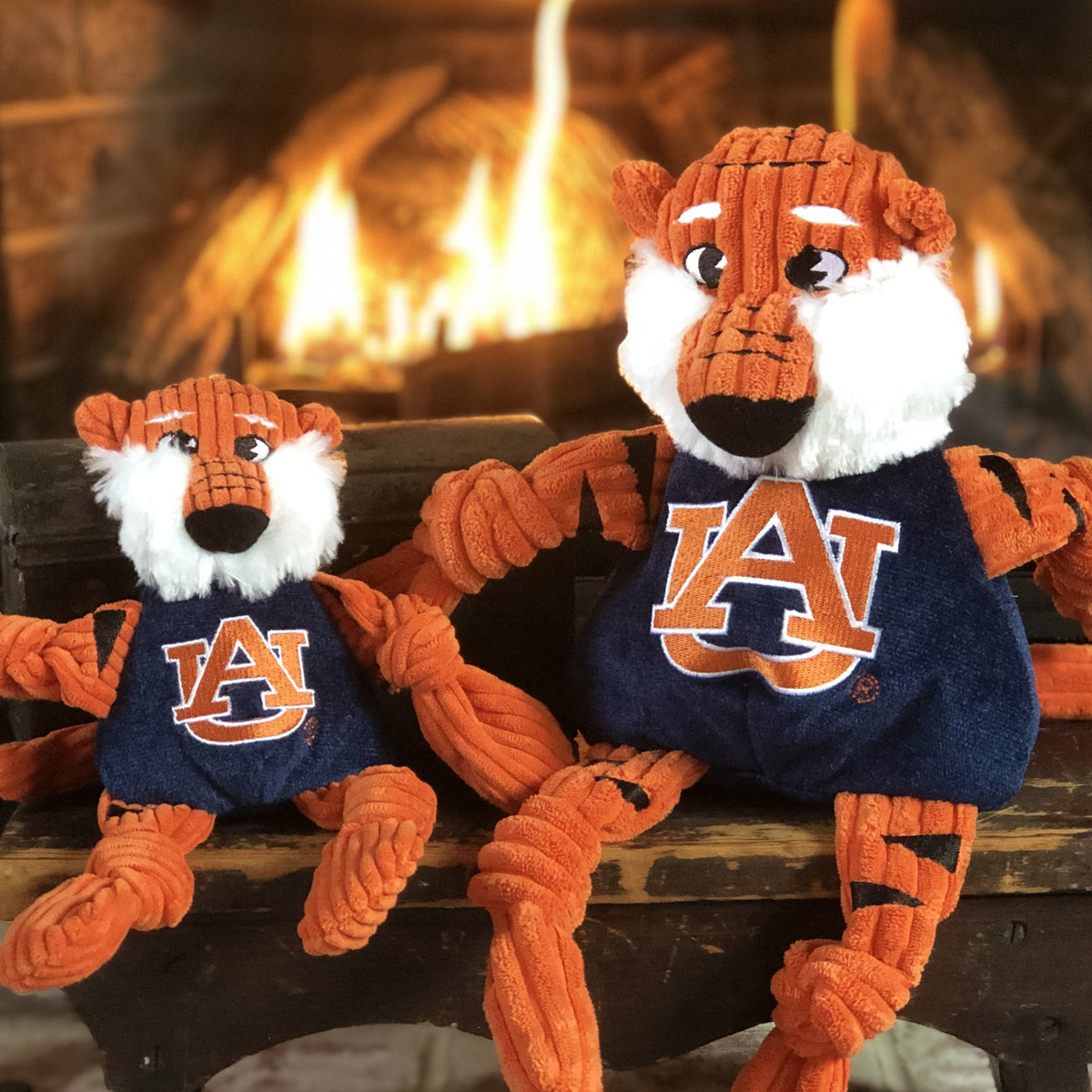 Aubie the Tiger Knottie dog toy in two sizes on a wooden bench in front of a lit fireplace