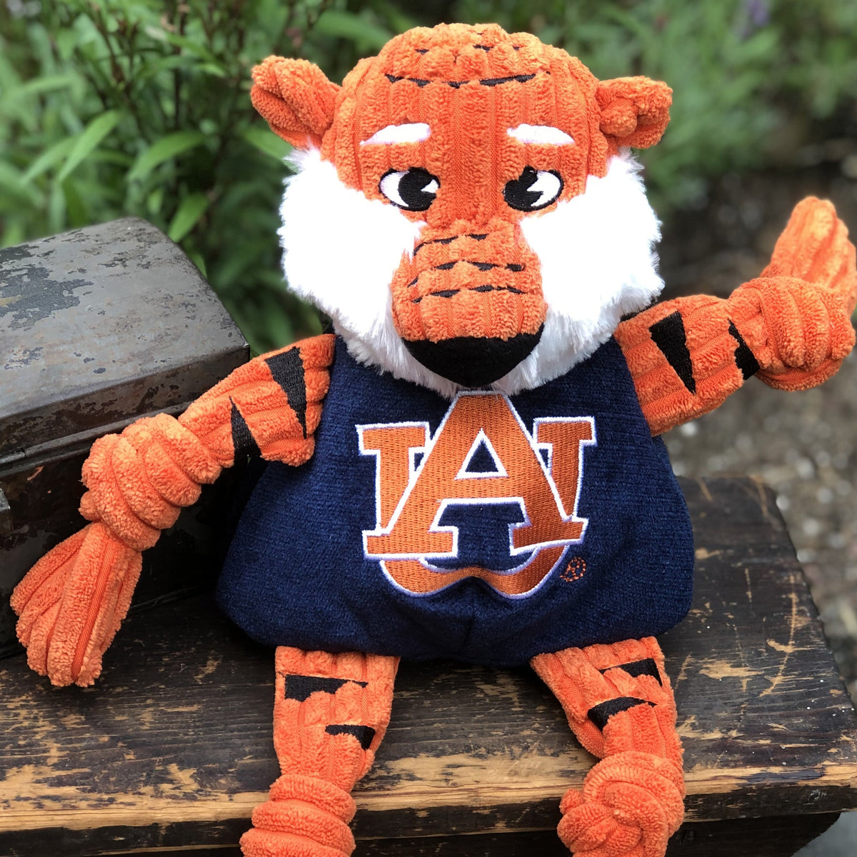 Aubie the Tiger Knottie dog toy sitting on a weathered wooden bench outdoors, one arm raised