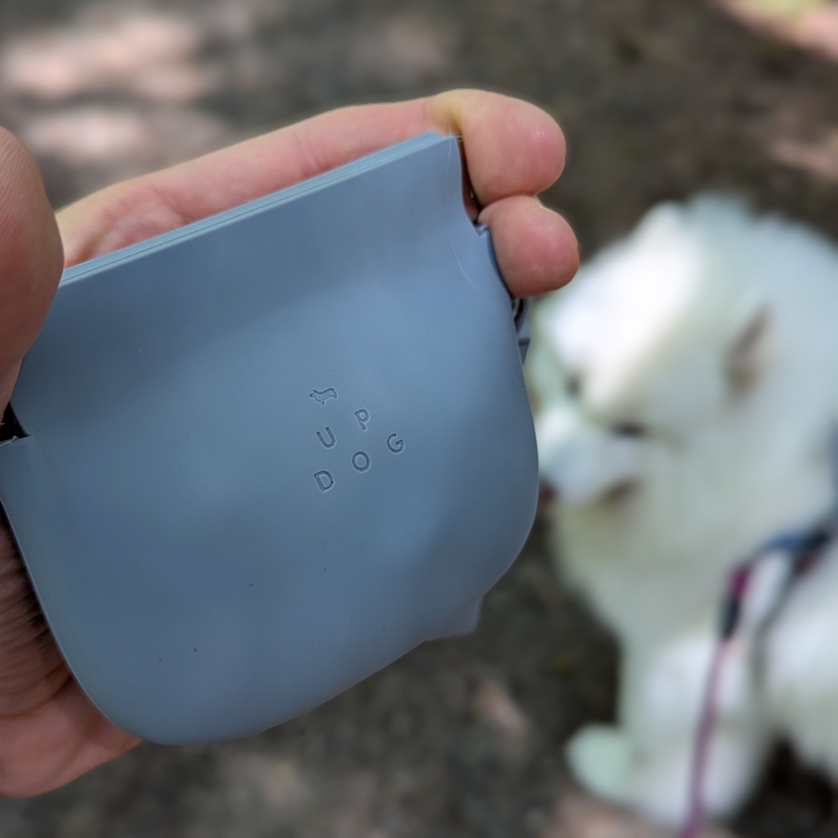 Up Dog One-Hand Treat Pouch in gray held closed in one hand on a walk, white fluffy dog waiting behind