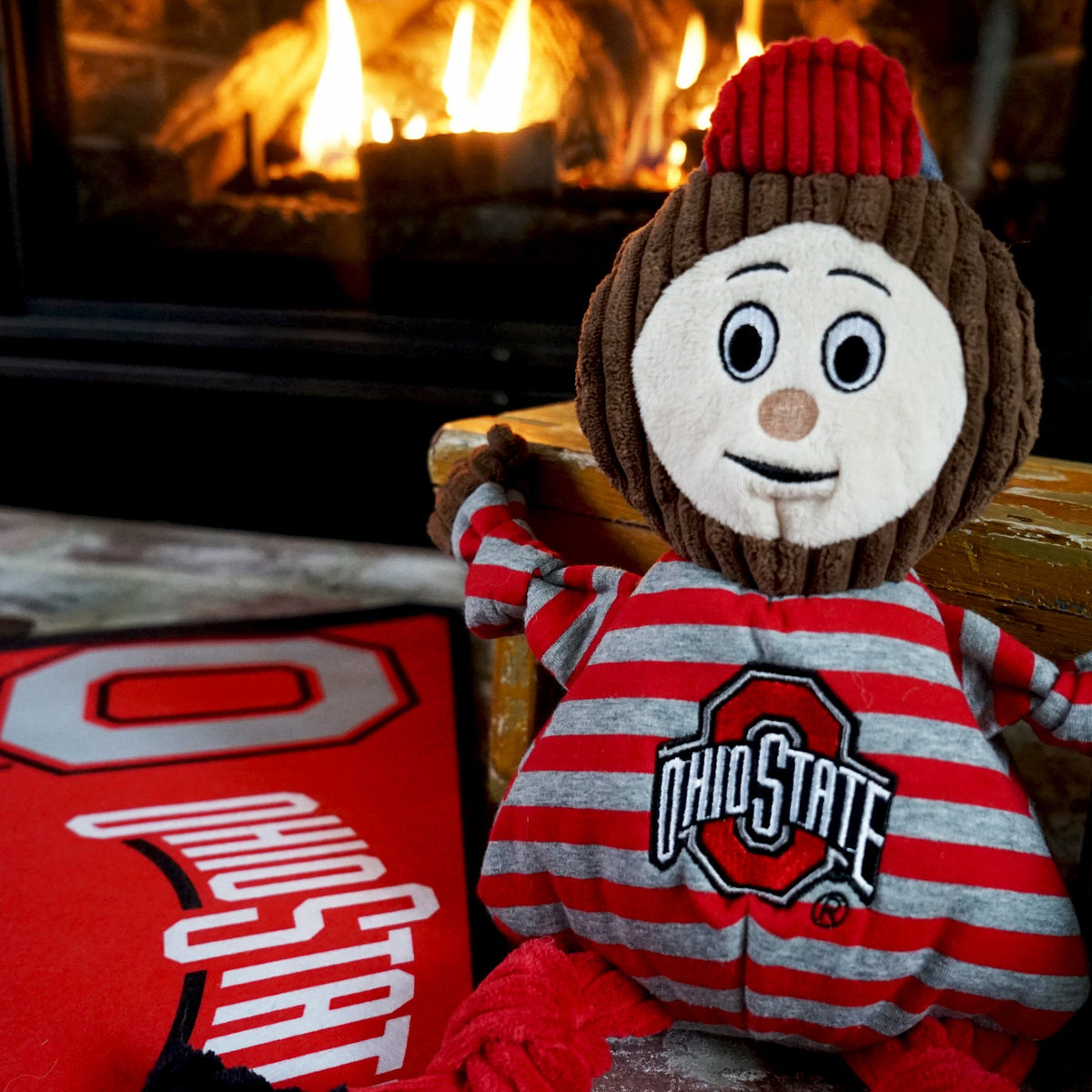 Brutus Buckeye Knottie dog toy sitting on a wooden bench by a lit fireplace, a red Ohio State banner beside it