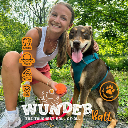 Smiling woman crouched beside a brown dog in a teal harness, holding an orange WUNDERball on a trail