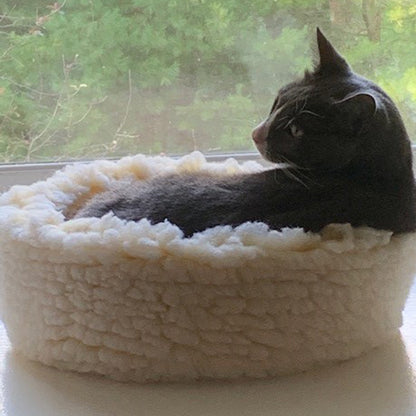 HuggleSnuggler fleece bed by a window, a gray and white cat lying inside and looking back over its shoulder