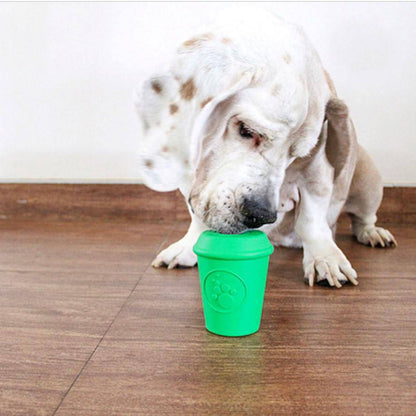 Basset hound licking the lid of the green Coffee Cup treat toy, standing upright on a wood floor