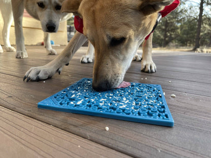 Jigsaw eMat lick mat in blue on a wooden deck, a tan dog licking it while a second dog watches